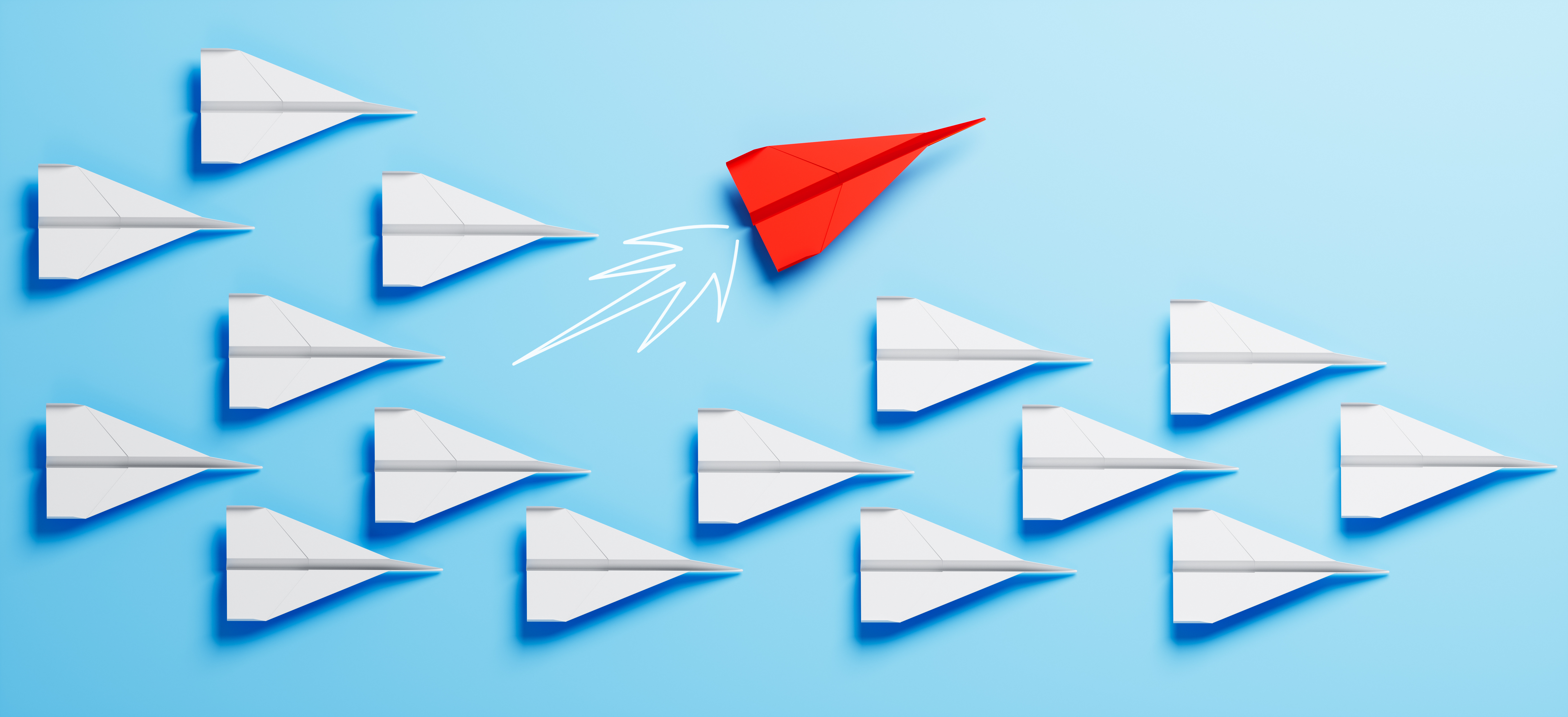 Das Bild zeigt viele weiße Papierflieger, die alle in dieselbe Richtung fliegen. Zwischen ihnen befindet sich ein einzelner roter Papierflieger, der in eine andere Richtung fliegt als die weißen. Der Hintergrund ist in einem kräftigen Türkis gehalten, wodurch die Papierflieger besonders gut zur Geltung kommen. Das Bild vermittelt den Eindruck von Individualität und Anderssein inmitten einer Gruppe.
