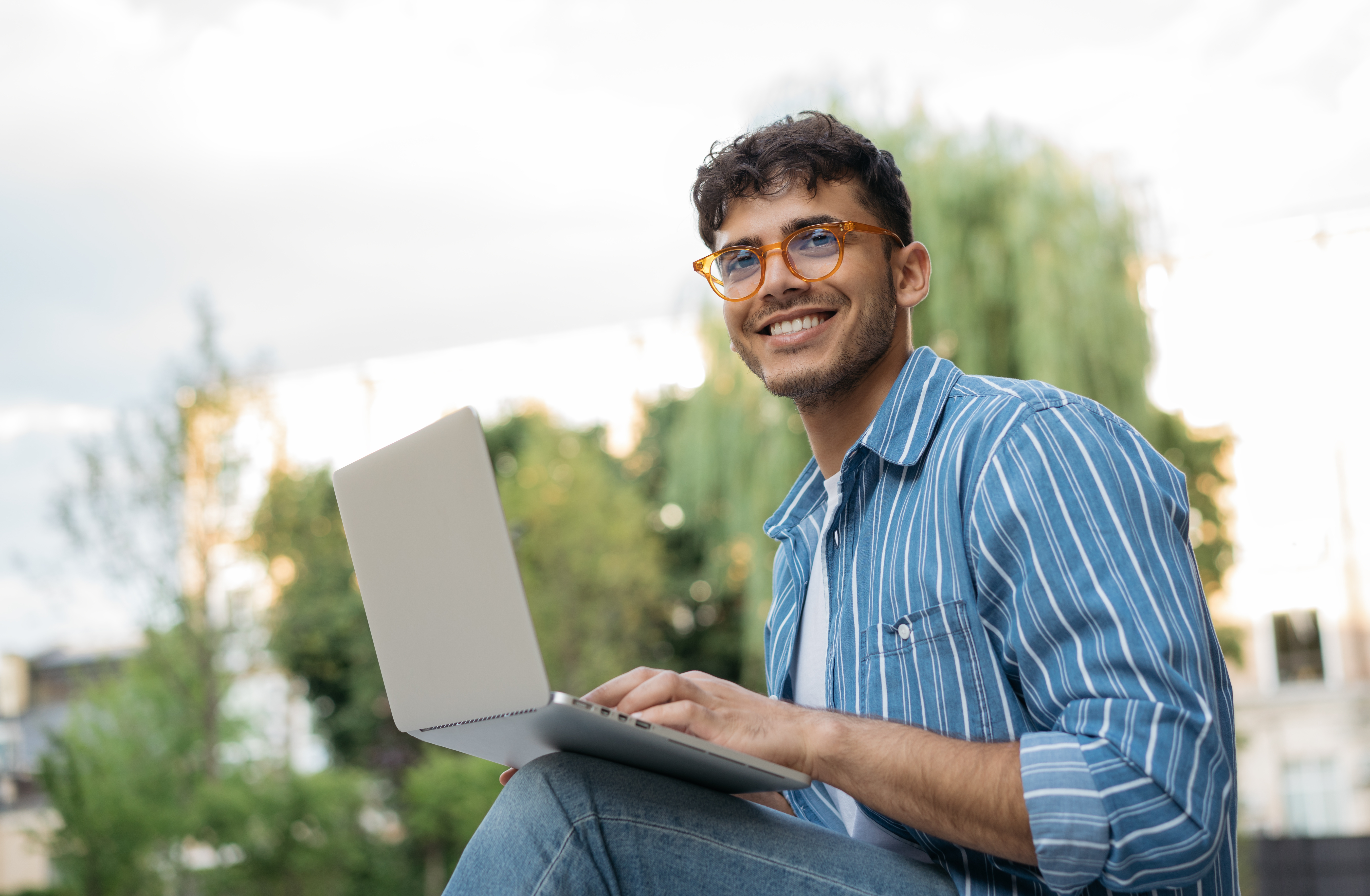 Ein Student sitzt in der Natur vor seinem Laptop. Er trägt eine Brille und ein blau gestreiftes Hemd über einem weißen T‑Shirt.