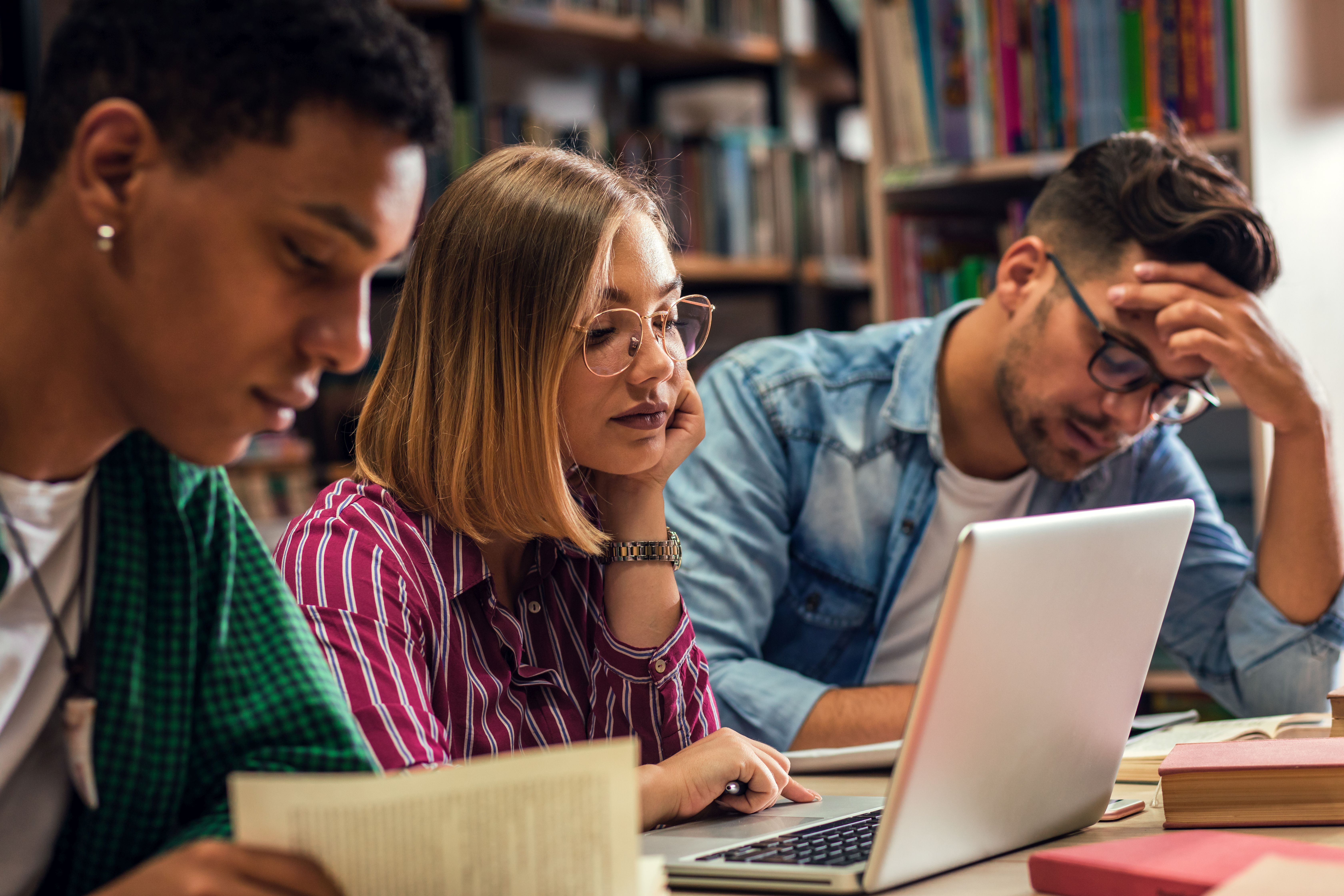 Zu sehen sind drei Personen, zwei Männer und eine Frau, die nebeneinander an einem Tisch sitzen. Ein Mann hat den Kopf auf den Arm aufgestützt. Die Frau sitzt in der Mitte und schaut in den offenen Laptop, der dritte Mann ist im Vordergrund zu sehen und liest in einem Buch.