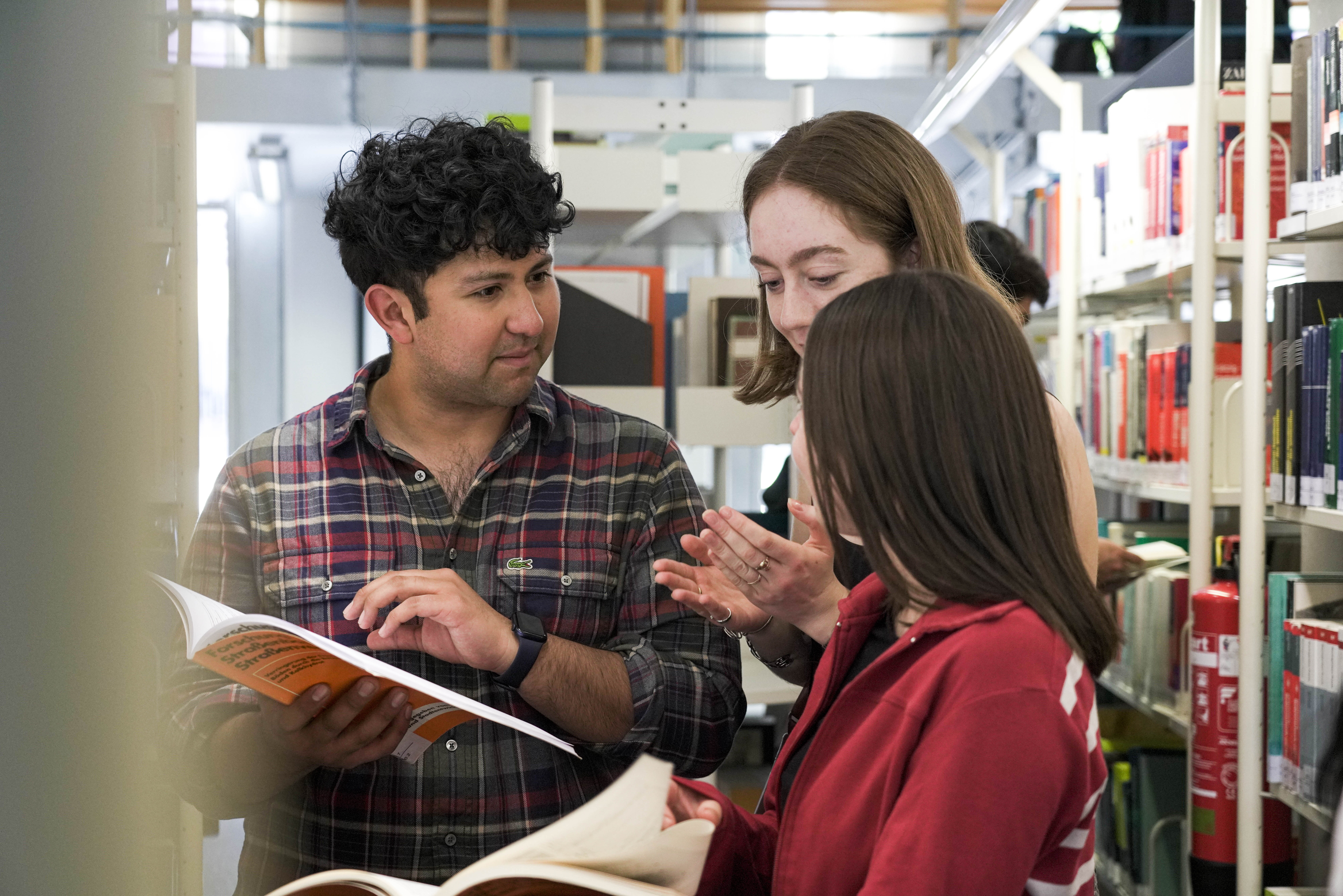 Drei Studierende stehen in Bibliothek und schauen in zwei Bücher