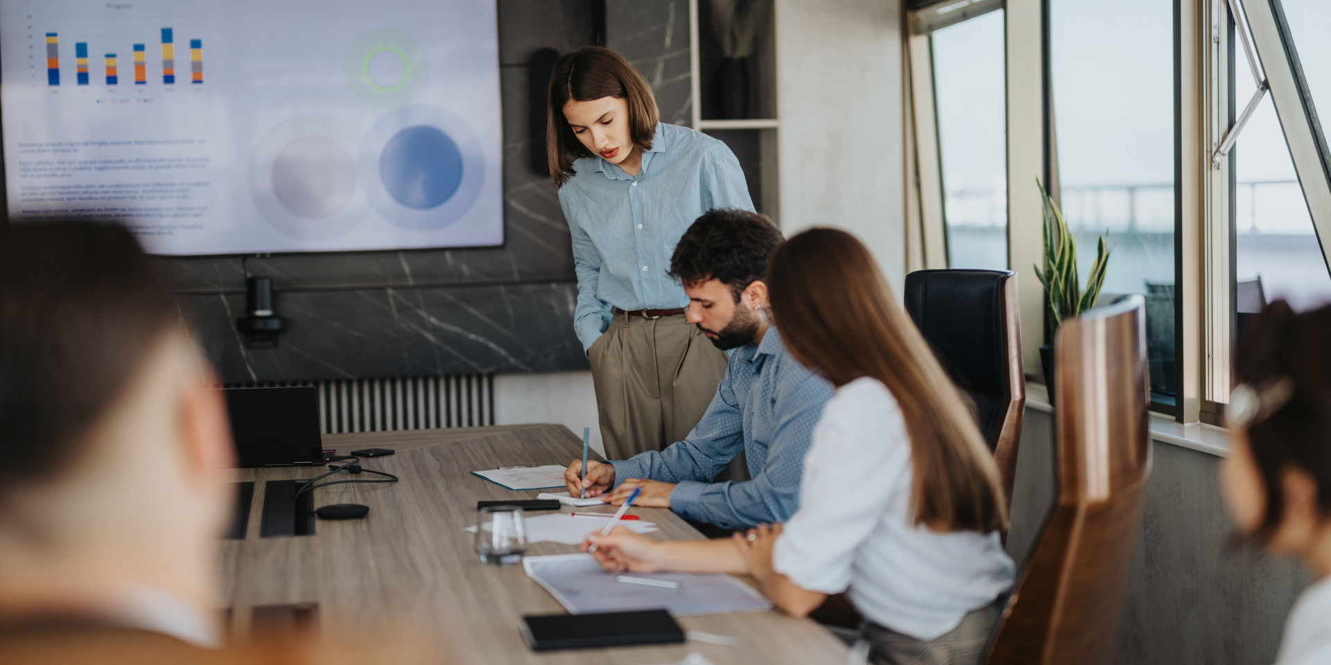 Moderner Besprechungsraum mit Berufstätigen im Smart-Casual-Look, die eine Strategiepräsentation mit Säulendiagrammen diskutieren.