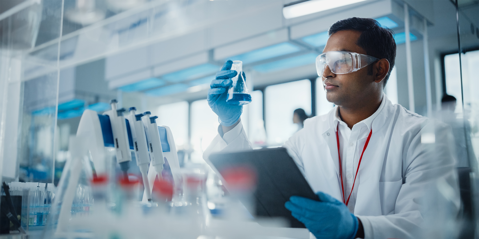 Männlicher Wissenschaftlers analysiert flüssige Biochemikalien in einem Erlenmeyerkolben und verwendet ein digitales Tablet in einem modernen Biotechnologielabor.