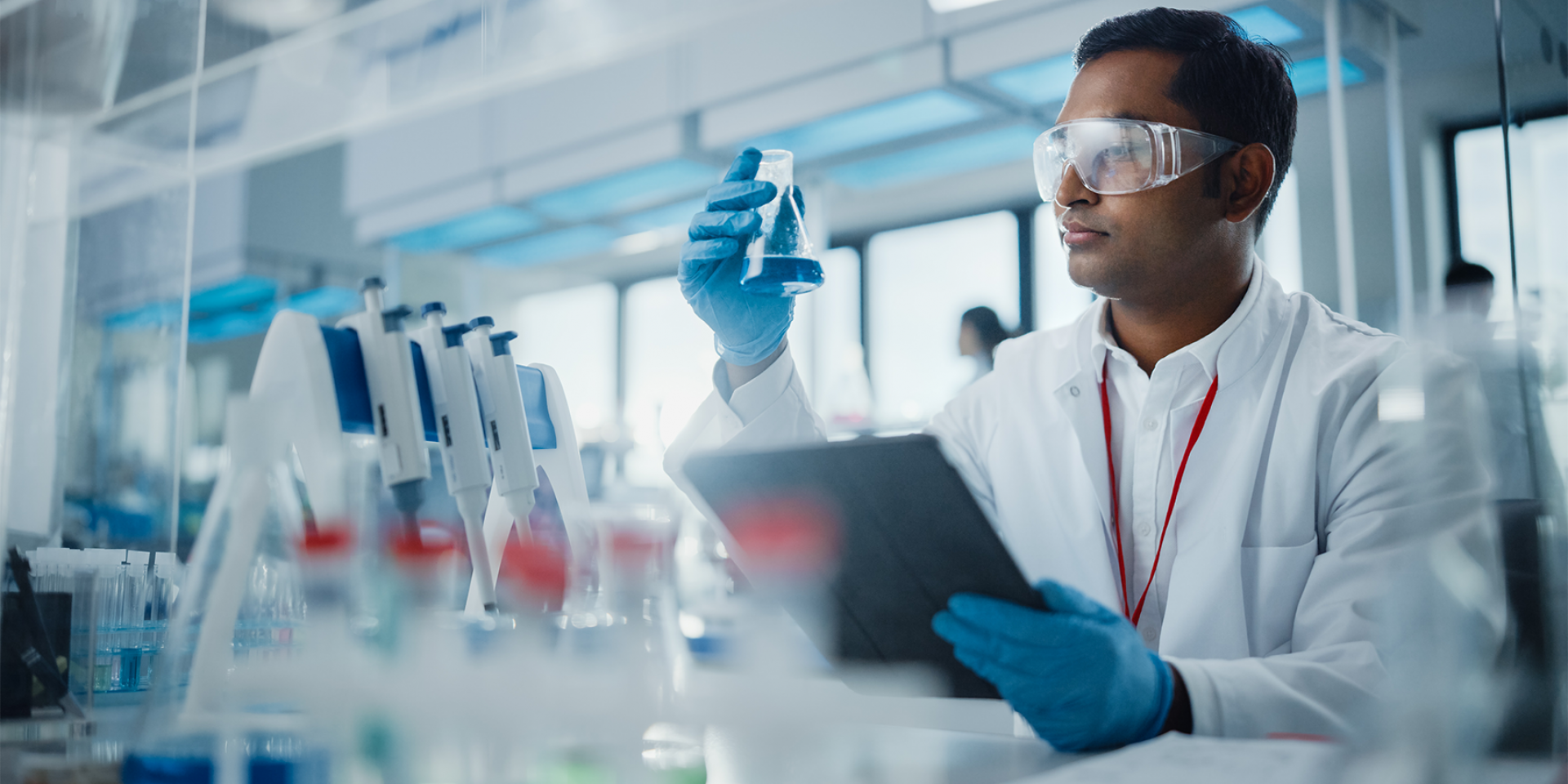 Männlicher Wissenschaftlers analysiert flüssige Biochemikalien in einem Erlenmeyerkolben und verwendet ein digitales Tablet in einem modernen Biotechnologielabor.