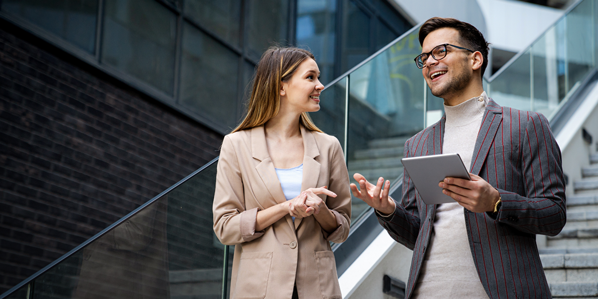 Eine Frau und ein Mann mit Tablet in der Hand, beide im Business Look, stehen auf einer Treppe in einem modernen Unternehmen und unterhalten sich,