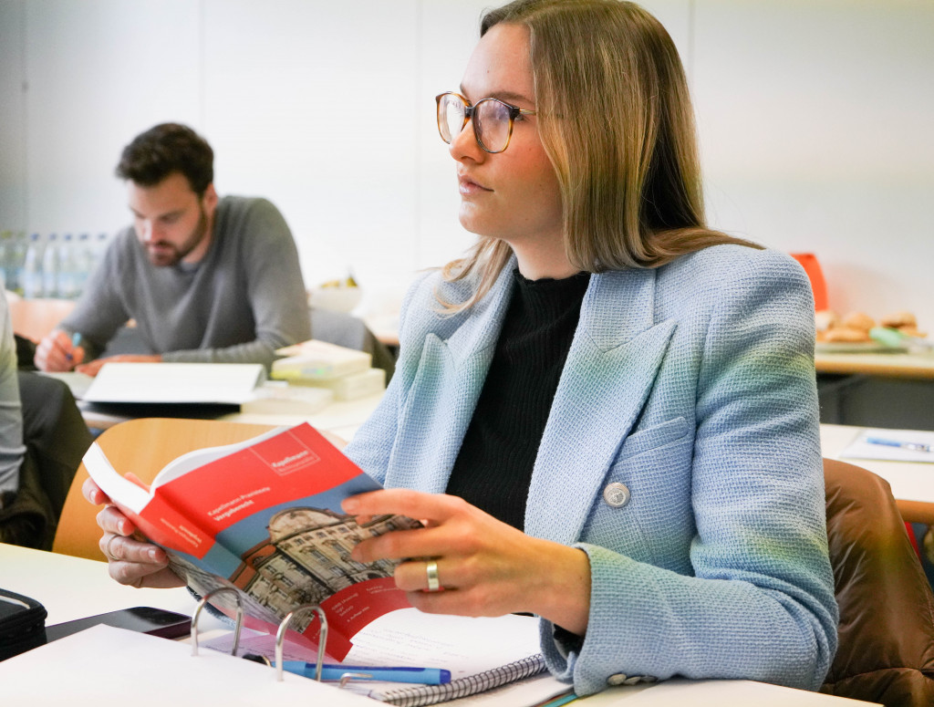 Berufsbegleitender Master Wirtschaftsrecht Junge Frau mit blauem Blazer sitzt in Vorlesung und hält Buch in der Hand