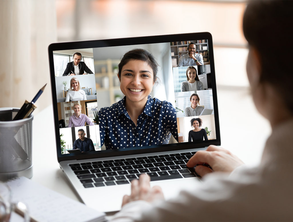Frau mit weißer Bluse sitzt vor einem Laptop am Schreibtisch und nimmt an einem Online-Meeting teil, dessen Teilnehmer auf dem Bildschirm zu sehen sind