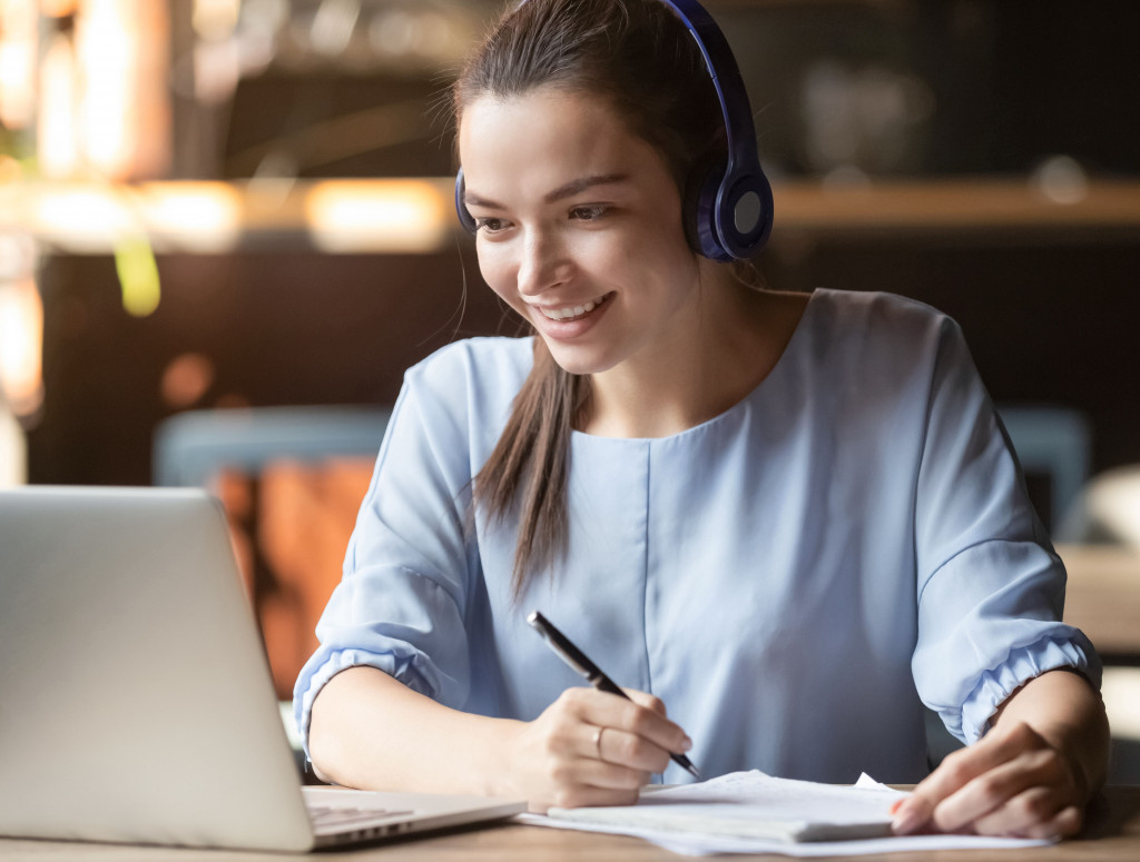 Auf dem Bild ist sitzt eine weibliche Studierende mit Headset vor einem Laptop und macht Notizen auf einem Papier wärend sie an einem Online-Seminar teilnimmt.