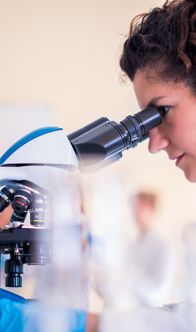 Junge Frau in weißem Kittel schaut durch ein Mikroskop in einem modernen Labor
