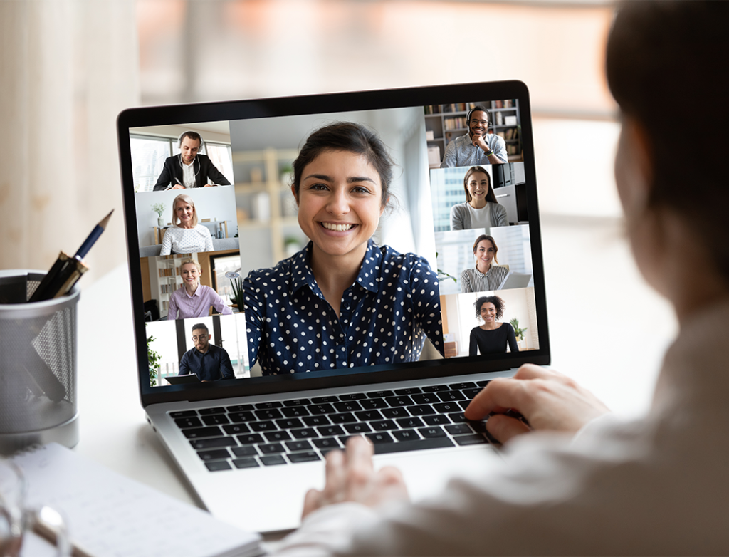 Frau mit weißer Bluse sitzt vor einem Laptop am Schreibtisch und nimmt an einem Online-Meeting teil, dessen Teilnehmer auf dem Bildschirm zu sehen sind