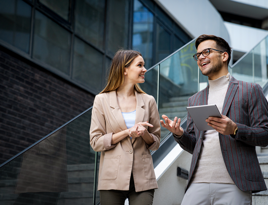 Eine Frau und ein Mann mit Tablet in der Hand, beide im Business Look, stehen auf einer Treppe in einem modernen Unternehmen und unterhalten sich,